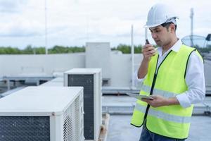 Asian maintenance engineer works on the roof of factory. contractor inspect compressor system and plans installation of air condition systems in construction. technology, online checking, mobile photo