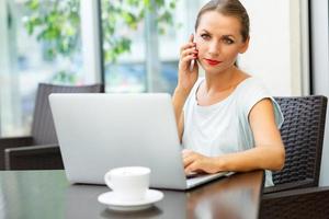 Young woman sitting in a cafe with a laptop and talking on the cell phone photo