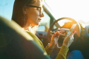 mujer en lentes usos un teléfono inteligente mientras conducción un coche a puesta de sol foto