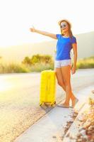 Young woman with a thumb up and yellow suitcase is traveling on the road hitchhiking photo