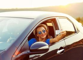 joven mujer en sombrero y Gafas de sol haciendo yo retrato sentado en el coche foto