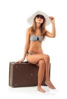 Full length portrait of a beautiful young woman posing in a bikini sitting on a suitcase on a white photo