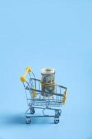 Small shopping trolley, cart with dollars banknotes roll inside isolated on blue background photo