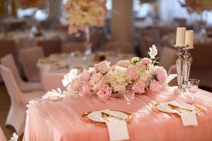 table decoration with pink tablecloth, crystal candlesticks with candles and white pink flowers in restaurant . stylish wedding day. setting table with gold plate, fork and knife photo