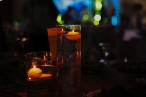 Banquet table decorated with burning candles in glass vases in restaurant hall. In the background party with silhouettes of people dancing on the dance floor with disco lights glowing searchlight photo