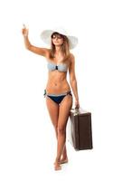 Full length portrait of a beautiful young woman posing in a bikini, hat and sunglasses with a suitcase in hand on white photo