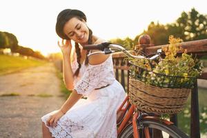 joven hermosa mujer sentado en su bicicleta con flores a puesta de sol foto