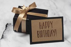negro regalo caja con un dorado cinta con un contento cumpleaños tarjeta en un blanco y negro mármol mesa. foto