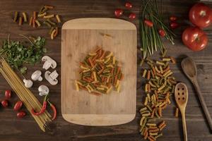 Flat lay of vegetables and pasta and a chopping table over a wooden table photo