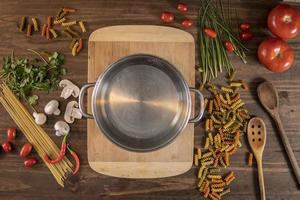 Flat lay of vegetables and pasta and a chopping board with a cacerola over a wooden table photo