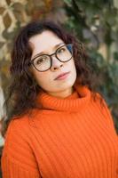 Autumn portrait of an attractive young woman in stylish glasses in a knitted fashionable orange sweater on background of leaves. Girl walks in city photo