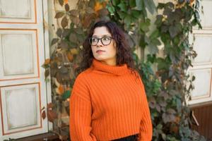 Stylish pensive thoughtful young woman wearing orange sweatshirt and eyeglasses. Portrait of smiling girl in trendy eyeglasses in city, copy space and empty space for advertising photo