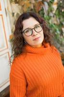 Autumn portrait of an attractive young woman in stylish glasses in a knitted fashionable orange sweater on background of leaves. Girl walks in city photo