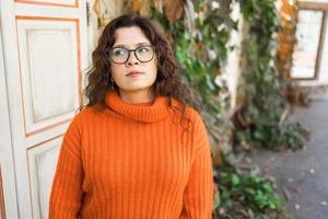 Stylish pensive thoughtful young woman wearing orange sweatshirt and eyeglasses. Portrait of smiling girl in trendy eyeglasses in city, copy space and empty space for advertising photo