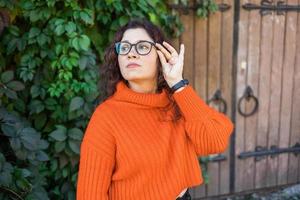 Portrait of carefree young woman smiling with big door background. Cheerful latin girl wearing eyeglasses in the city. Happy brunette woman with curly hair and spectacles smiling. photo