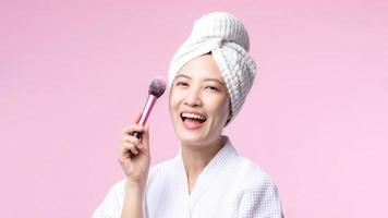 Portrait happy young asian woman with natural make up face holding cosmetic skin powder blusher isolated on pink background. Female apply skincare brush treatment. beauty product, cosmetology concept. photo