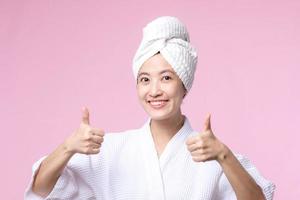 Beautiful young asian woman happy smile face with hand or finger gesture signs in bathrobe and shower towel showing empty space isolated on pink background. photo