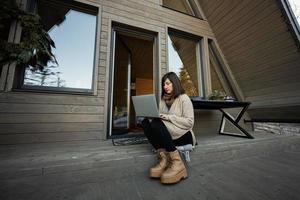 Remote work and escaping to nature concept. Woman works on laptop against tiny cabin house.