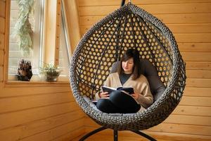 Remote work and escaping to nature concept. Woman sit in egg chair swing and read book in wooden tiny cabin house. photo