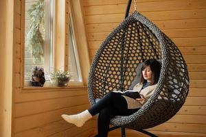 Remote work and escaping to nature concept. Woman sit in egg chair swing and read book in wooden tiny cabin house. photo