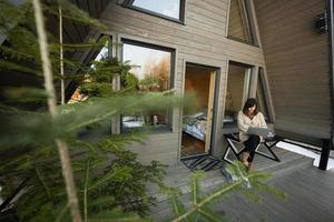 Remote work and escaping to nature concept. Woman works on laptop against tiny cabin house. photo