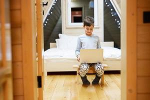 chico en pijama sentar con ordenador portátil a de madera cabina hogar. concepto de infancia, ocio actividad, felicidad. foto