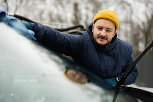 el hombre limpia el parabrisas de un vehículo suv americano con un paño de microfibra después de lavarlo en clima frío. foto