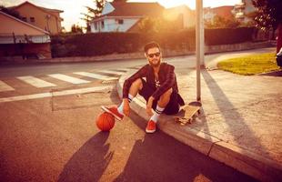 joven elegante hombre con un baloncesto y patineta sentado en un ciudad calle a puesta de sol ligero foto