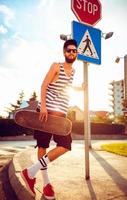 elegante hombre en Gafas de sol con un patineta en un calle en el ciudad a puesta de sol ligero foto