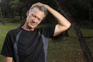 Mature Man outside at a park sitting on a tree stump stretching out his neck for pain relief photo