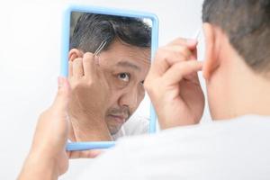 asiático medio hombre mira en el espejo y usos pinzas a arrancar su gris pelo foto