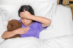 asiático niño niña enfermedad acostado en cama y poner mano en frente a cheque temperatura, foto