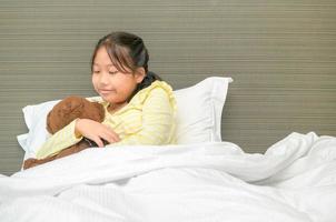dulce pequeño niña es abrazando un osito de peluche oso, mirando a oso y sonriente mientras acostado en su cama foto