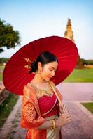 Beautiful Thai girl in traditional dress costume red umbrella as Thai temple where is the public place, Thai Woman in Traditional Costume of Thailand. photo