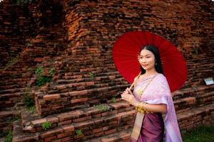 hermosa tailandés niña en tradicional vestir disfraz rojo paraguas como tailandés templo dónde es el público lugar, tailandés mujer en tradicional disfraz de tailandia foto