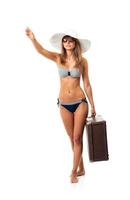 Full length portrait of a beautiful young woman posing in a bikini, hat and sunglasses with a suitcase in hand on white photo