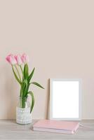 Home interior with decorative elements. Layout with a white frame and pink tulips in a vase and a pink notebook on the table on a light beige background photo