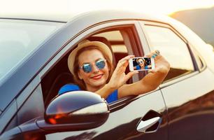 joven mujer en sombrero y Gafas de sol haciendo yo retrato sentado en el coche foto