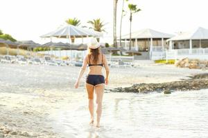 mujer en un sombrero y traje de baño caminando en el playa en un caliente verano día - ver desde el espalda foto