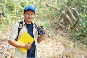 Asian man explorer wears blue cap, holds binocular in forest to survey botanical plants and creatures wildlife. Concept, nature exploration. Ecology and Environment. photo