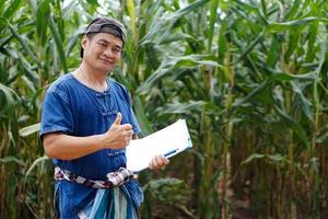 Portrait of Asian man farmer is at maize garden,hold paper clipboard, Thumbs up. Concept , Agricultural study and research. Organic farming. photo