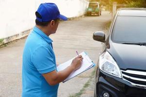 Asian man mechanic wears blue cap and blue shirt, holds paper notepad, checking and evaluating broken car condition.Concept,  claim for accident insurance. Roadside assistance. Outdoor car service. photo