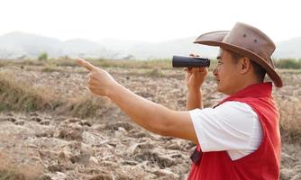 Asian man explorer wears hat, red vest shirt, holds binocular to explore boundary , points to something. Concept, land property exploration. Long vision distance, zoom for clearing  scene. Observation photo