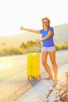 Young woman with a thumb up and yellow suitcase is traveling on the road hitchhiking photo
