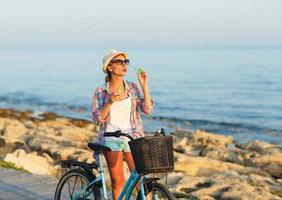 Carefree woman with bicycle standing on a wooden path at the sea and blow bubbles photo