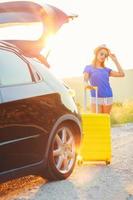 Woman with a yellow suitcase standing near the trunk of a car parked on the roadside photo