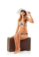 Full length portrait of a beautiful young woman posing in a bikini sitting on a suitcase on a white photo