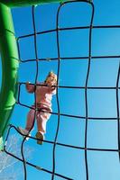 contento pequeño activo niña subido en un cuerda web en un patio de recreo en contra un brillante azul cielo en un soleado día foto