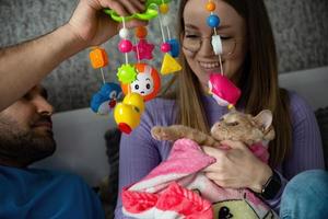 A young couple on the bed hugs their cat wrapped in a baby blanket and plays with her with a baby rattle. Conscious refusal of a couple to have children, treatment of pets as children. photo