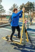 A retired woman with a backpack on her shoulders on an active walk outdoors is engaged in exercise equipment in the park. Senior woman taking care of her health photo